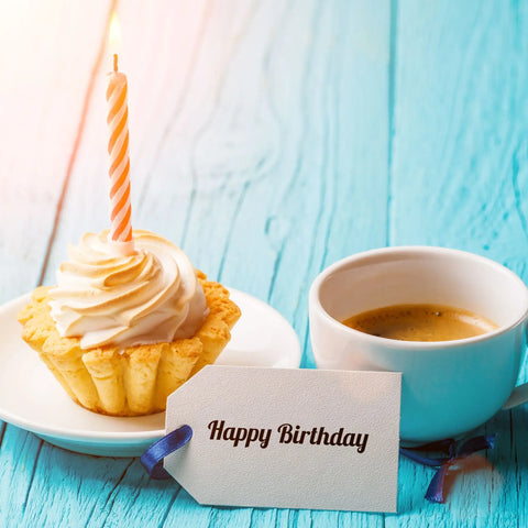 A birthday cupcake with a candle and a cup of coffee sitting on a blue table.