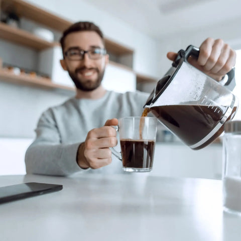 A guy pouring coffee from a drip coffee maker into his coffee mug.