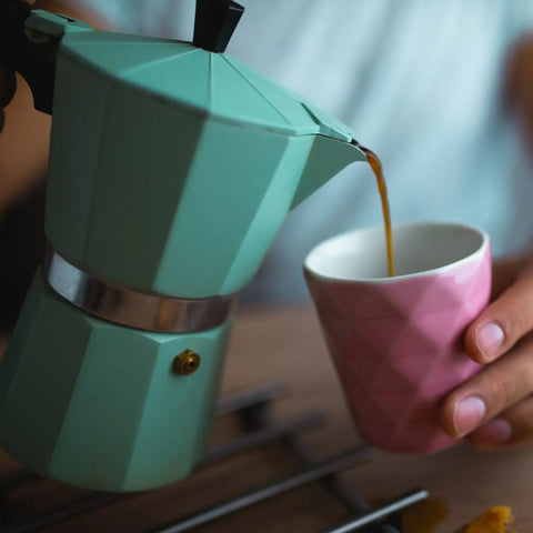 A person pouring coffee into a pink mug from a baby blue Moka pot.