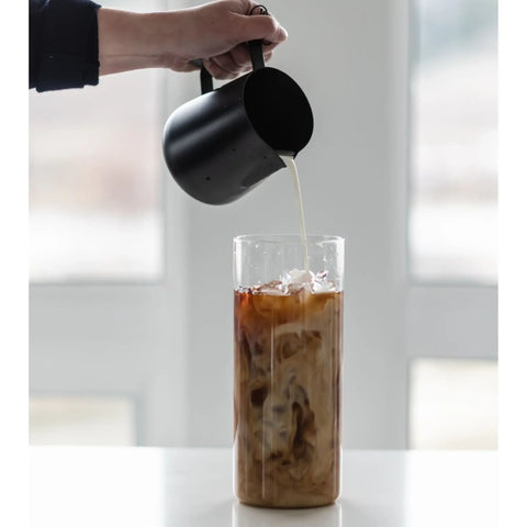 A person pouring cream from a pitcher into a large glass of cold brew.