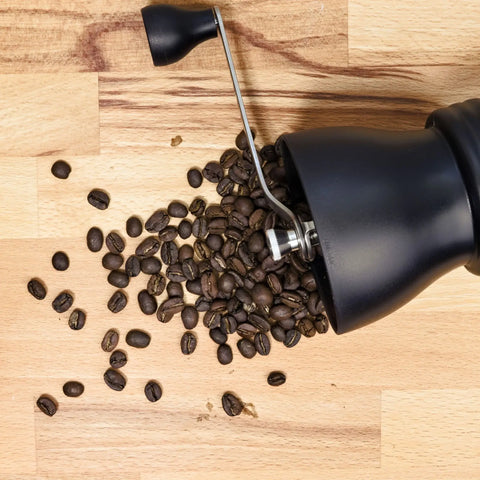 A manual coffee grinder on a wooden counter with beans spilling out.